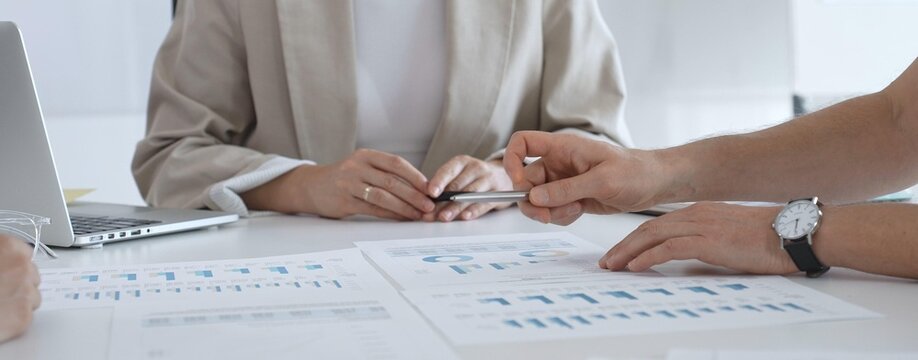Professional businesspeople collaborating on financial analysis, female executive gesturing toward charts while presenting strategic insights to team members in corporate workspace