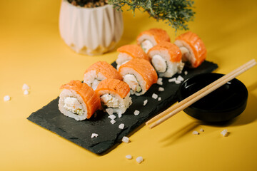 Sushi rolls with salmon on a slate plate and chopsticks placed on a yellow background