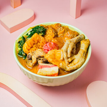 A bowl of spicy seblak with noodles, chicken feet, fish cake, and vegetables, served in rich orange broth. Captured on a pink background, showcasing Indonesia&rsquo;s famous street food in vibrant style.