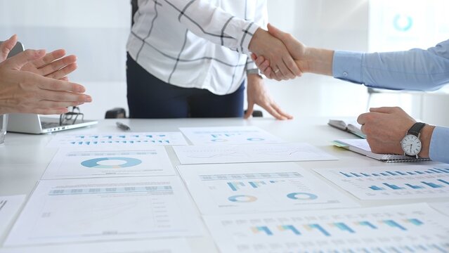 Business partnership success concept with handshake in modern office, financial charts, graphs and laptop on desk representing teamwork, agreement and corporate growth