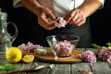 A chef carefully removes petals from fresh blossoms, preparing them for a dish. Surrounding ingredients include lemon, salt, and herbs, enhancing the cooking process