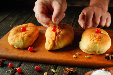A baker carefully places a bright viburnum on top of each freshly baked sweet bun. The warm, golden pastries sit on a wooden board surrounded by spices, creating a cozy atmosphere