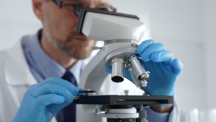 Close up of male scientist in white coat and blue gloves adjusting microscope, conducting pharmaceutical research in modern laboratory. Medicine and science concept