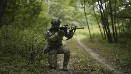 Camouflaged and masked army soldier with AR15 carbine on a war recon mission. Kneeling on the grass to aim the weapon. Military recon