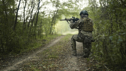 Camouflaged and masked army soldier with AR15 carbine on a recon mission. Kneeling on the grass to aim the weapon. Military recon