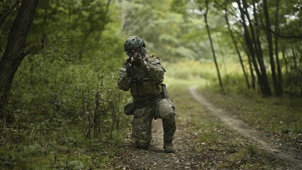 Camouflaged and masked army soldier with AR15 carbine on a war recon mission. Kneeling to aim. Military recon
