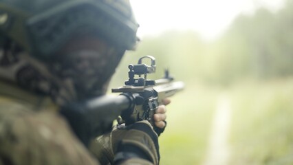 Camouflaged and masked army soldier with AR15 carbine on a recon mission. Standing on a grass field and aiming. Military recon