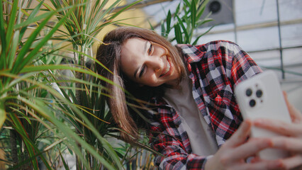 Young woman takes a selfie in a greenhouse surrounded by lush greenery during daylight hours to...
