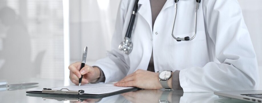 Female doctor in white coat with stethoscope writing patient notes on clipboard, completing medical records in clinic office. Medicine and health care - Powered by Adobe