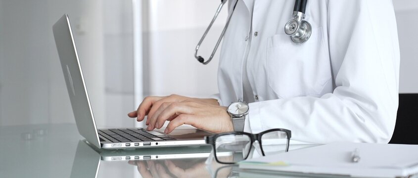 Woman doctor wearing white coat and stethoscope, typing on laptop in clinic office, using modern medical technology. Medicine, healthcare and science concept