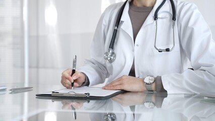 Female doctor in white coat with stethoscope writing patient notes on clipboard, completing medical...