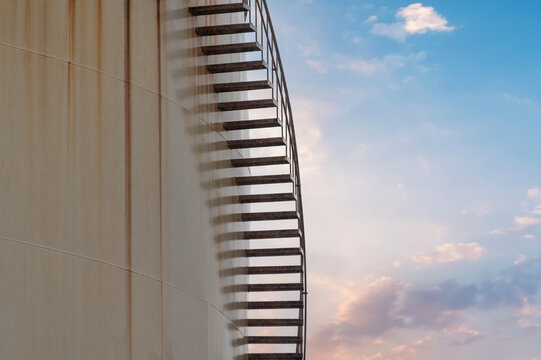 Large Industrial tanks for oil storage with metal stairs on the side and a sunset sky in the background. This elegant and slender staircase climbs up the side of an oil or petroleum storage tank.