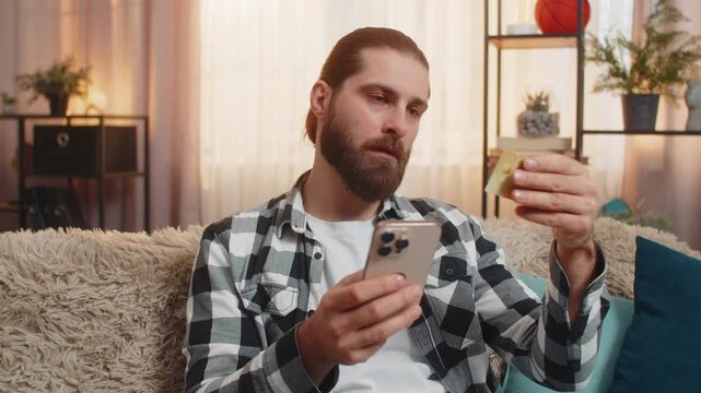Young man sits at home with phone and credit card, finalizing online store purchase while verifying total. Caucasian guy on sofa presses ok, nodding confidently over successful payment transaction