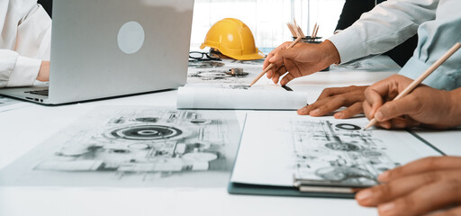 A dynamic team collaborates over engineering plans in a modern office setting. The scene captures hands working on documents with a laptop and a helmet, highlighting creativity. SACTR