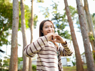 Fototapeta premium Smiling young woman making heart shape with her hands in forest setting, surrounded by tall trees and natural light, expressing joy and love for nature