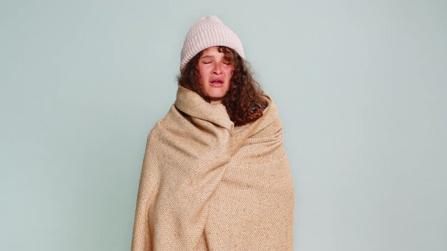 Woman wearing hat, wrapped in warm blanket, inhaling deeply and coughing due to cold, flu illness symptoms. Girl on gray background shows signs of feeling sick, unwell, discomfort respiratory health.