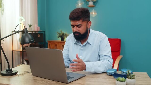 Indian man at home sees laptop error rubs forehead sighs upset virus glitch causing serious trouble. Arabian guy at table frustrated seeks quick fix hopes system recovers rapidly stress growing today.