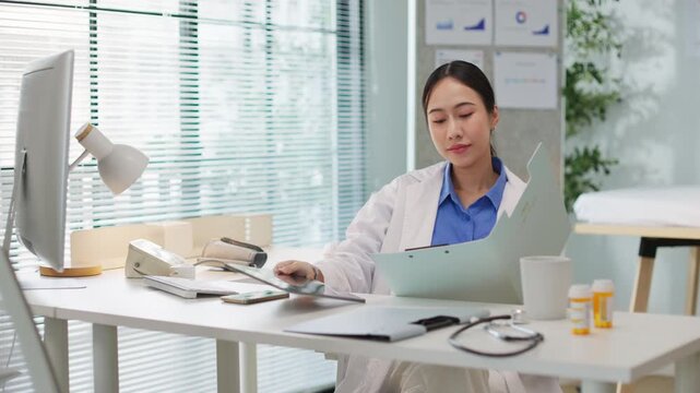 Asian female doctor in white coat sitting at desk in clinical office hospital, typing on computer, reviewing patient records, manage medical data, analyzing healthcare test results planning treatment.