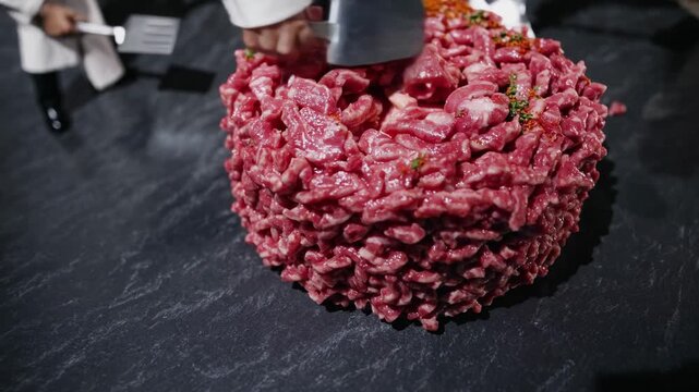 Chefs are using metal spatulas to serve a large portion of steak tartare from a giant mound of freshly ground beef seasoned with spices on a dark gray table