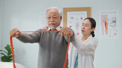 Asian female physiotherapist helps an elderly man perform resistance band exercises for physical therapy in a modern clinic, promoting rehabilitation, strength training, and active aging support.