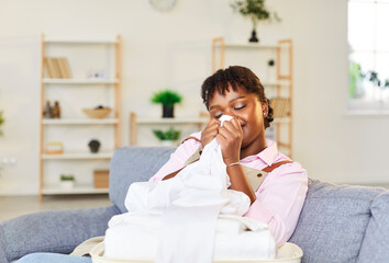Young smiling African American woman sitting on couch in living room, smelling fresh clean clothes in laundry after washing with new detergent gel. Joyful girl sniffing scent of washing powder. 