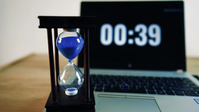 Hourglass with blue sand in front of a laptop showing a countdown timer