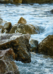 A vertical shot of a beautiful wave breaking against coastal rocks creating a dramatic splash of white foam on the brown stones