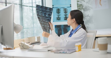 Asian female doctor examines MRI film in clinic, reviewing patient data and medical imaging for diagnosis. Professional healthcare, radiology analysis, spine or neurological check, treatment planning.