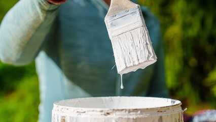 Obraz premium A person dips a paint brush in white paint from a matching bucket. This image symbolizes home improvement and DIY projects, perfect for illustrating renovation and artistic applications.