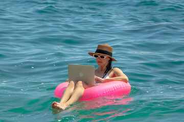 Remote work digital nomad ocean. Woman working on a laptop while floating in the blue sea on a pink inflatable ring enjoying summer freedom.