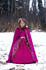 Cute toddler girl in a beautiful velvet royal princess outfit with a cape and a long dress. Child is standing in the snowy forest, Halloween costume. © Saeedatun