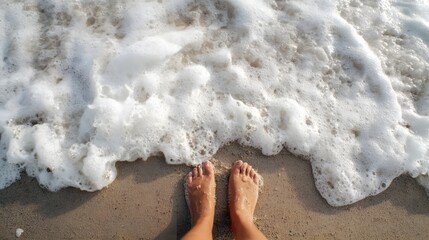 Bare feet stand on wet sand as sea foam washes ashore in an overhead view