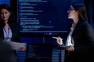 Two female engineers one Asian and one Indian discuss data with a male colleague in a command center. They are analyzing AI code for a cybersecurity system.