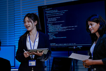 Two female engineers one Asian and one Indian discuss data with a male colleague in a command center. They are analyzing AI code for a cybersecurity system.