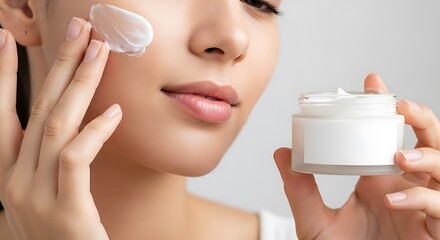 Young woman applying white facial cream to her cheek from a small jar, focusing on skincare routine.