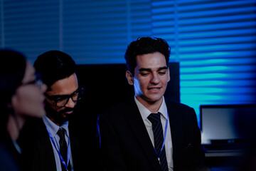 A cybersecurity team with a Caucasian engineer and an Indian analyst in a dark control room. They are working late, analyzing a system threat or AI data.