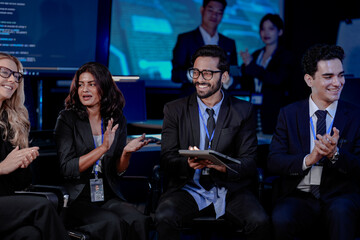 Obraz premium An Indian man with a tablet asks a question during a large team briefing. His diverse colleagues, including Indian and Caucasian women and men, listen in a high-tech conference room.