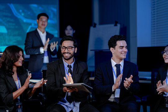 A large, diverse team of business professionals in a group meeting. An Indian man with a tablet leads the discussion with Asian and Caucasian colleagues in a high-tech office.