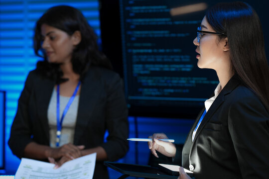 An Asian businesswoman with glasses leads a discussion, holding a tablet and stylus. Her Indian female colleague smiles and holds a document in a modern tech office.