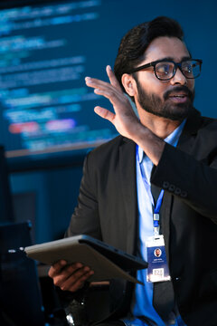 An Indian male software engineer or data scientist holds a tablet in a command center. He is asking a question about the AI code or cybersecurity data shown on the large screen.