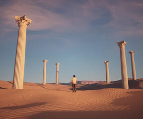 Man walking through desert ruins with ancient columns under a bright blue sky during sunset