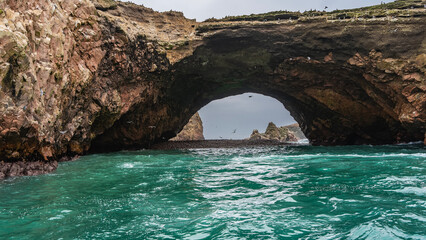 Natural stone arch in the rock. Seabirds are sitting on a cliff, flying in the sky. Turquoise ocean. The pebble beach. Peru. Paracas. The Ballestas Islands