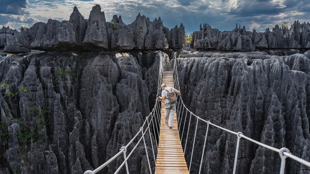 A man with a backpack and safety gear walks across the suspension bridge over the gorge. He holds onto the rope railing, looking down into the abyss. There are steep cliffs with sharp peaks ahead.