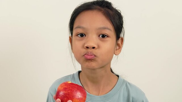 A video shows a girl standing holding a red apple and biting it bit by bit, almost halfway through. With different poses and perspectives, she smiles brightly as she enjoys eating her favourite fruit.