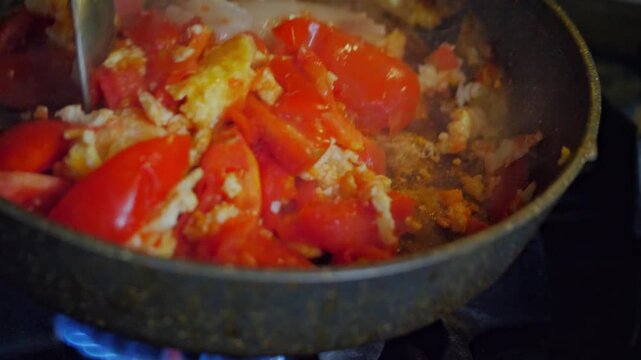 An active and savory mood is captured by a close-up of a spoon stirring scrambled eggs and chopped tomatoes in a hot frying pan. This shot is ideal for depicting home cooking, breakfast