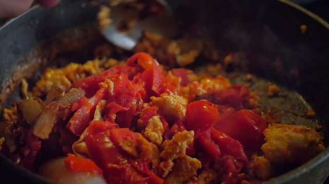 An active and savory mood is captured by a close-up of a spoon stirring scrambled eggs and chopped tomatoes in a hot frying pan. This shot is ideal for depicting home cooking, breakfast