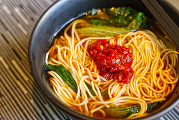 close-up of a dark bowl of hot noodle soup topped with a large dollop of vibrant red chili paste and green vegetables. 