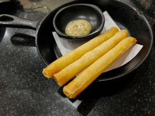 Three golden-brown fried spring rolls or cheese sticks served in a small black cast iron skillet with a side of creamy dipping sauce