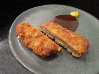 Two pieces of deep-fried, breaded meat cutlet (likely Beef or Pork Katsu), one sliced open, served on a modern grey-green plate. The dish is complemented by a thick brown sauce and a dollop of mustard