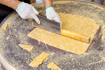 A close-up of a street vendor or baker cutting a large sheet of traditional yellow peanut brittle or sesame snack with a large knife on a round metal table.